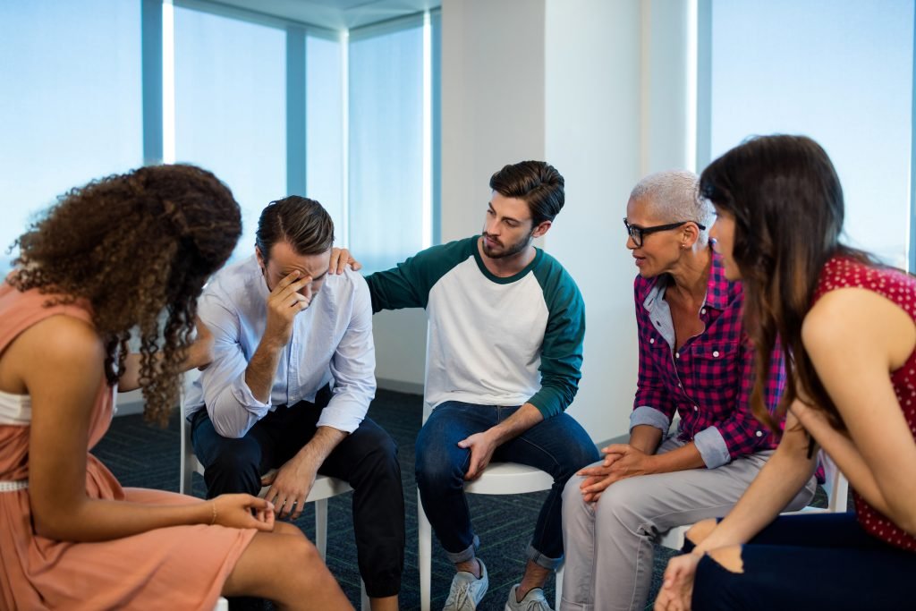 Grupo de cinco personas sentadas en círculo en una sala, brindando apoyo a un hombre que parece estar angustiado.